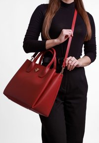 Rote Lederhandtasche mit strukturierter Form, doppelten Griffen und einem abnehmbaren Schultergurt. Mit einem goldenen Logo-Detail auf der Vorderseite.