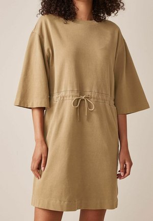 Kobieta ubrana w luźną, sięgającą kolan beżową sukienkę z rękawami do łokci i ściągaczem wiązanym w talii.