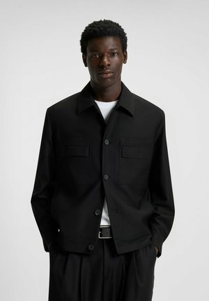 Black jacket with a collared neckline, two front pockets, and button closures. Worn over a white shirt, paired with black pants. Smooth fabric.