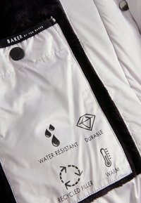 Weißes Stoffetikett mit schwarzen Akzenten, versehen mit Symbolen und Text: „Wasserabweisend“, „Langlebig“, „Recycelte Füllung“ und „Warm“.