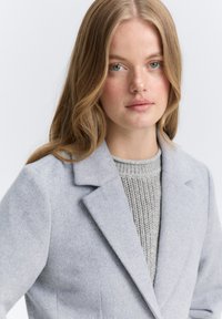 Lichtgrijze wolblend blazer met notch-lapels, grote voorzakken en gestructureerde silhoutte, over een textuur grijze gebreide trui.