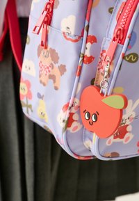 Sac à dos violet avec impressions d’animaux de dessin animé et de fleurs, fermetures éclair rouges et un charm en forme de pomme rouge avec un visage souriant sur la tirette.