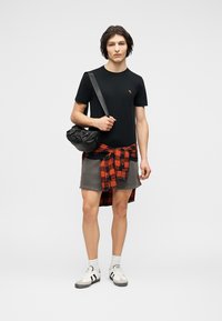 Νεαρός άνδρας με μαύρο μπλουζάκι, γκρι σορτς, κόκκινο πουκάμισο με καρό δεμένο στη μέση, λευκά αθλητικά παπούτσια και μαύρη τσάντα χιαστί, μπροστά σε απλό φόντο.