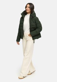 Veste matelassée verte avec un col montant et une fermeture éclair, associée à un pantalon large crème. La veste présente un design matelassé et une capuche réglable.