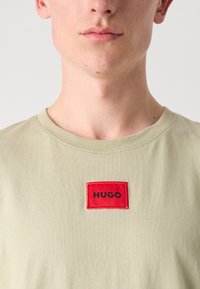 Tricou din bumbac verde deschis, cu o petrecere rectangulară roșie proeminentă pe care este brodat textul "HUGO" în negru pe față. Guler rotund.