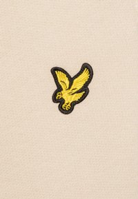 Tissu en tricot beige avec un logo d'aigle brodé jaune entouré de noir, doté d'une texture fine et d'une finition lisse.
