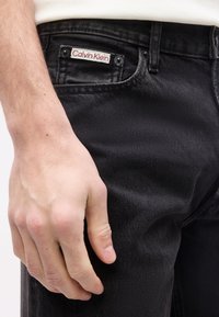 Κοντινό πλάνο ενός χεριού που ακουμπά δίπλα στην τσέπη μαύρου τζιν με μια μικρή ετικέτα Calvin Klein στην άκρη της τσέπης.