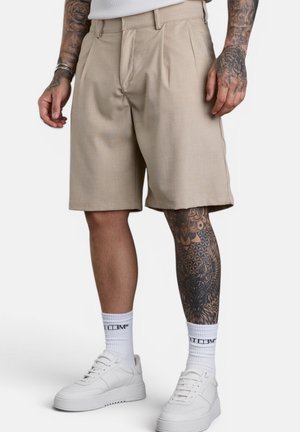Mann trägt beige knielange Shorts, weiße Socken, weiße Sneakers und hat tätowierte Arme und rechtes Bein auf weißem Hintergrund.