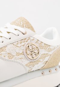 Hvid sneaker med guldglimmer, snørebåndsmønster på siden, guldlogo-detalje og sølvnitter på sålen. Snørebåndene er hvide.