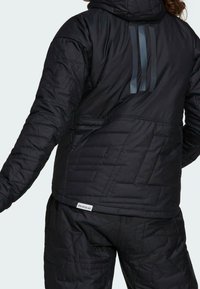 Sort quiltet jakke med hætte, med teksturerede mønstre og reflekterende striber på ryggen. Mærket lyder "PRIMEBLUE" ved kanten.