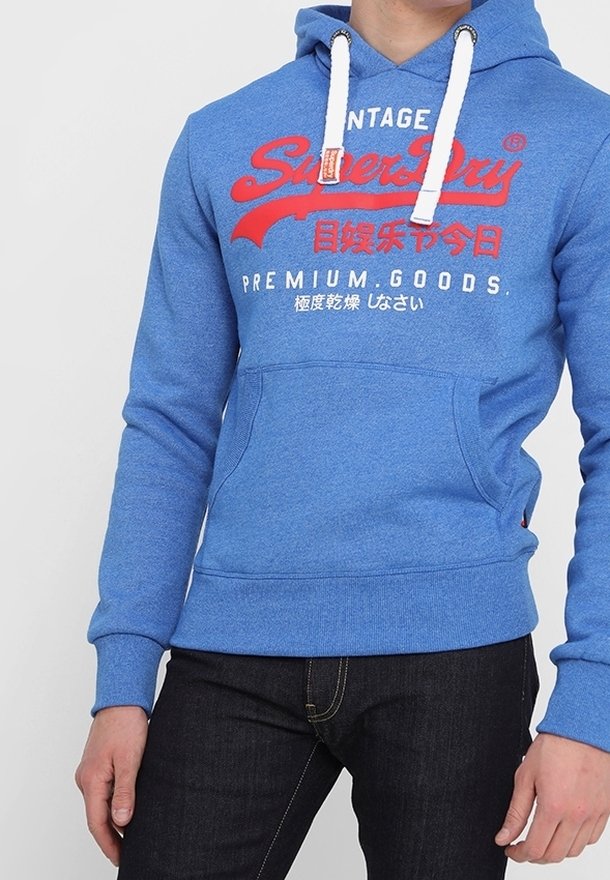 Sweat à capuche bleu avec cordons blancs, texte imprimé en rouge et blanc en anglais et en japonais, porté avec un jean foncé.