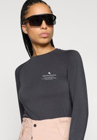 Mustapaita, jossa on pitkät hihat ja raglan-hihat, joissa on valkoinen logo. Yhdistettynä vaaleanpunaisiin housuihin. Mallilla on tummat aurinkolasit ja suorat letitetyt hiukset.