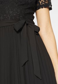 Jupe plissée noire avec un nœud à la taille, associée à un haut noir en dentelle à manches courtes sur le haut du corps d'une personne.