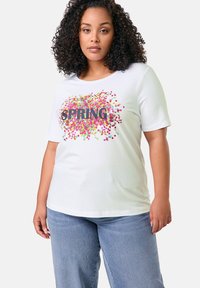 Weißes Baumwoll-T-Shirt mit buntem "SPRING"-Text und Konfetti-Muster. Kurze Ärmel und lockere Passform, kombiniert mit hellblauen Jeans.