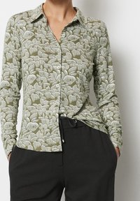 Camicia verde con bottoni e maniche lunghe, caratterizzata da un motivo di foglie bianche e un tessuto testurizzato. Abbinata a pantaloni neri.