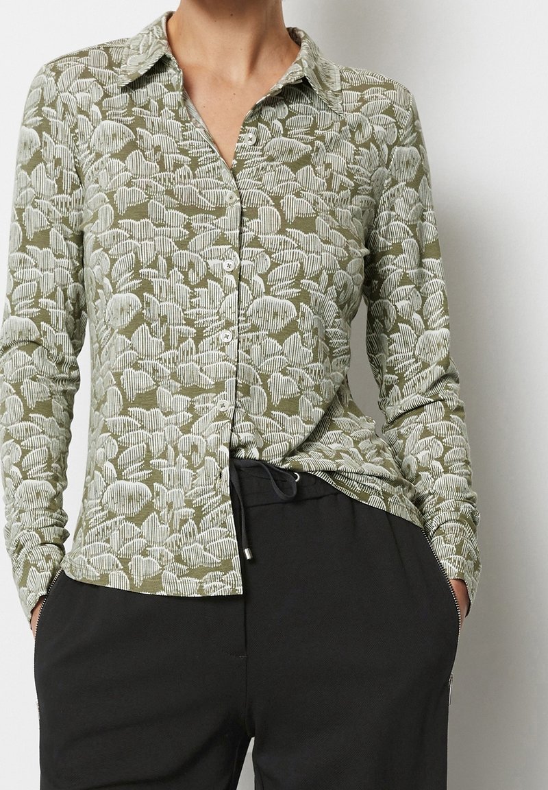 Camicia verde con bottoni e maniche lunghe, caratterizzata da un motivo di foglie bianche e un tessuto testurizzato. Abbinata a pantaloni neri.