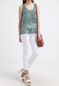 Top senza maniche verde e bianco con motivo a foglia di palma, abbinato a jeans skinny bianchi e sandali platform bianchi. Borsa a tracolla marrone.