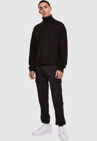 Giovane uomo che indossa un maglione a collo alto in nero a trecce, pantaloni cargo neri e sneakers bianche, in piedi di fronte a uno sfondo neutro.