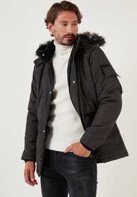 Black insulated jacket with a fur-lined hood, zippered front, and multiple pockets. Worn over a white ribbed turtleneck and black distressed jeans.