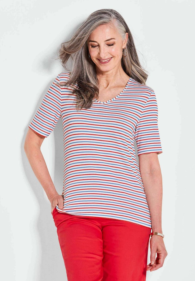 Gestreiftes Kurzarm-T-Shirt in Weiß, Rot und Blau. Mit einem V-Ausschnitt und weichem Stoff. Kombiniert mit roten Hosen.