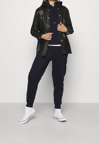 Chaqueta de cuero negro sobre una sudadera con capucha azul marino, combinada con pantalones deportivos azul marino y zapatillas blancas de caña alta. Textura suave, ajuste casual, herrajes mínimos.