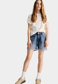 Camiseta blanca de algodón con mangas remangadas, pantalones cortos de mezclilla azul con puños y zapatos tipo slip-on color tostado. Modelo de pie con un fondo neutro.