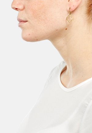 Boucles d'oreilles en or avec un pendentif en forme de trèfle. Texture lisse, affichant un design simple mais élégant. Portées avec un haut blanc à col rond.