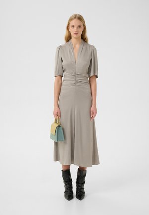 Graues knielanges Kleid mit Puffärmel, V-Ausschnitt und gerafften Details. Das Model hält eine zweifarbige Handtasche mit gelben und türkisfarbenen Akzenten. Schwarze knielange Stiefel.