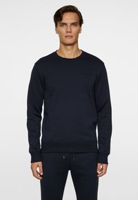 Mörkblå sweatshirt tillverkad av mjukt tyg, med rundad halsringning, långa ärmar och en subtil präglad logotyp på bröstet.