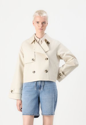 Beige kort dobbeltradet jakke med store knapper, vid krage og lange ermer, kombinert med blå denimshorts. Glatt tekstur.
