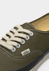 Πράσινο παπούτσι Vans ελιάς με λευκές τρύπες για κορδόνια, παχιά κρεμ κορδόνια, λευκή ραφή και ετικέτα με το λογότυπο Vans στο πλάι.