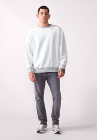 Pullover grigio chiaro con vestibilità morbida, polsini e orlo a costine, abbinato a jeans grigio scuro e sneakers metalliche.