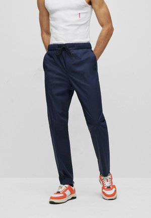 Homme portant un pantalon à cordon bleu marine, un débardeur blanc avec le logo rouge "HUGO" et des baskets orange et blanches, debout les mains dans les poches.