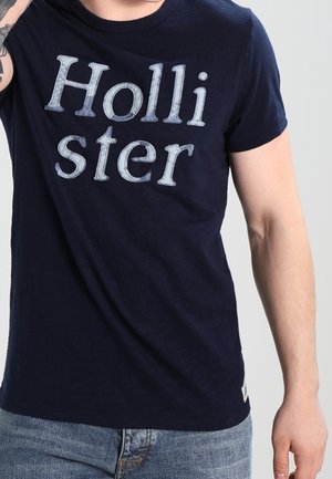 Osoba ubrana w granatową koszulkę Hollister i jasnoniebieskie dżinsy na jednolitym jasnym tle.