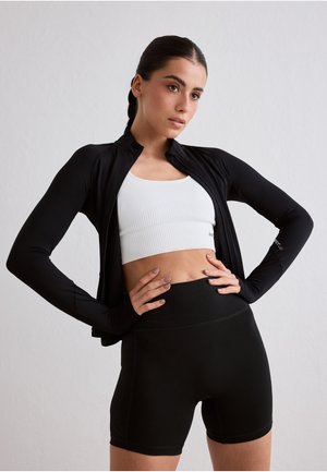 Kobieta o ciemnych, splecionych warkoczach, ubrana w biały sportowy biustonosz, czarną kurtkę zapinaną na zamek z otworami na kciuki oraz czarne, wysokowaistowe krótki spodenki rowerowe.