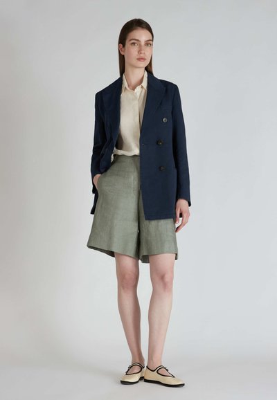 Marineblauwe dubbelbreasted blazer over een crèmekleurig shirt, gecombineerd met lichtgroene shorts en crèmekleurige schoenen met zwarte accenten. Zachte texturen, nauwkeurige pasvorm.