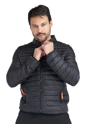 Un homme ajuste le col d’une veste noire matelassée avec des tirettes de fermeture éclair orange, sur fond blanc.