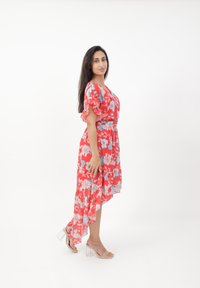 Robe fleurie rouge avec des manches courtes à volants, un ourlet asymétrique et des accents bleus. Associée à des sandales à talons transparents sur un fond neutre.