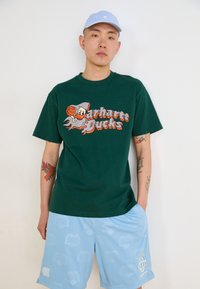 Grün/kurzärmliges T-Shirt mit "Carhartt Bucks" Grafik, kombiniert mit hellblauen Shorts mit einem dezenten Herzmuster. Hellblappe Mütze.