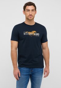 Tamsiai mėlyna trumpomis Rankovėmis marškinėliai su grafiniu dizainu, vaizduojančiu arklį ir užrašu "Mustang Jeans" kontrastingomis spalvomis priekyje.