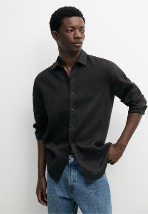 Junger Mann trägt ein schwarzes Hemd mit Knöpfen und hochgekrempelten Ärmeln sowie blaue Jeans, steht mit einer Hand in der Hosentasche und schaut zur Seite.