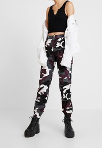 Crop top noir côtelé, veste blanche, et pantalon camouflage à motifs gris, noir et bordeaux. Des bottines noires épaisses à lacets complètent la tenue.
