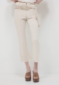 Jeans beige a cropped con orlo grezzo, dotati di una cintura decorativa con accenti in oro intrecciato e frange, abbinati a sandali platform marroni.