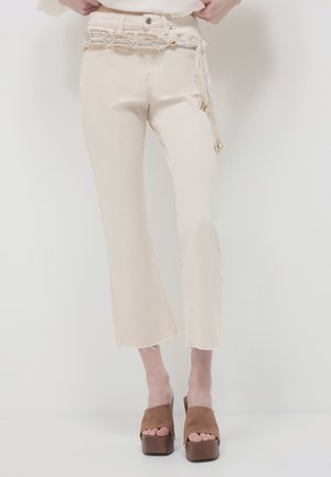 Jeans beige a cropped con orlo grezzo, dotati di una cintura decorativa con accenti in oro intrecciato e frange, abbinati a sandali platform marroni.