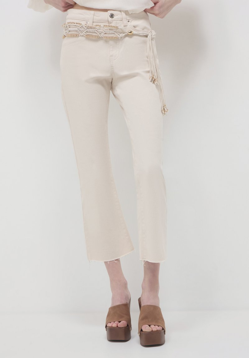 Jeans beige a cropped con orlo grezzo, dotati di una cintura decorativa con accenti in oro intrecciato e frange, abbinati a sandali platform marroni.