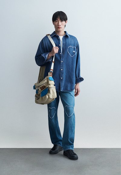 Jeune homme portant une chemise en denim et un jean avec des patchs en forme de cœur, portant un sac à bandoulière beige et bleu, debout sur un sol gris contre un mur blanc.