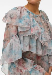 Blusa con stampa floreale presenta volant traslucidi e stratificati in tonalità pastello di rosa, blu e grigio, realizzata in un tessuto leggero e arioso con maniche arricciate.