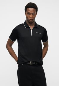 Νεαρός άνδρας φοράει μαύρο κοντομάνικο πόλο με λευκή ρίγα και φερμουάρ μπροστά, με τα χέρια πίσω από την πλάτη, μπροστά σε απλό φωτεινό φόντο.