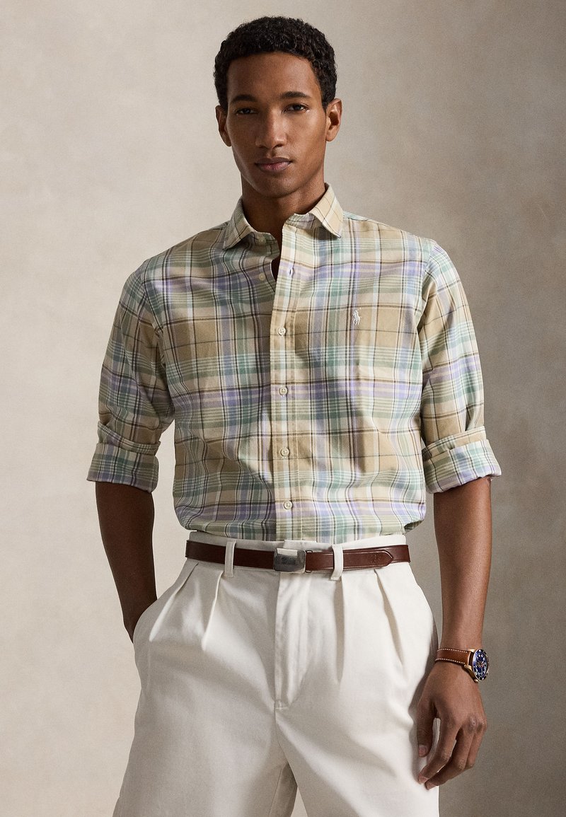 Un homme porte une chemise beige à carreaux pastel boutonnée, manches retroussées, rentrée dans un pantalon blanc plissé avec ceinture marron et montre.