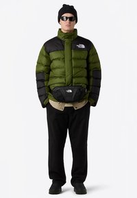 Green and black puffer jacket with a zip front, paired with a black fanny pack, beige shirt, black pants, and black shoes, worn with sunglasses.